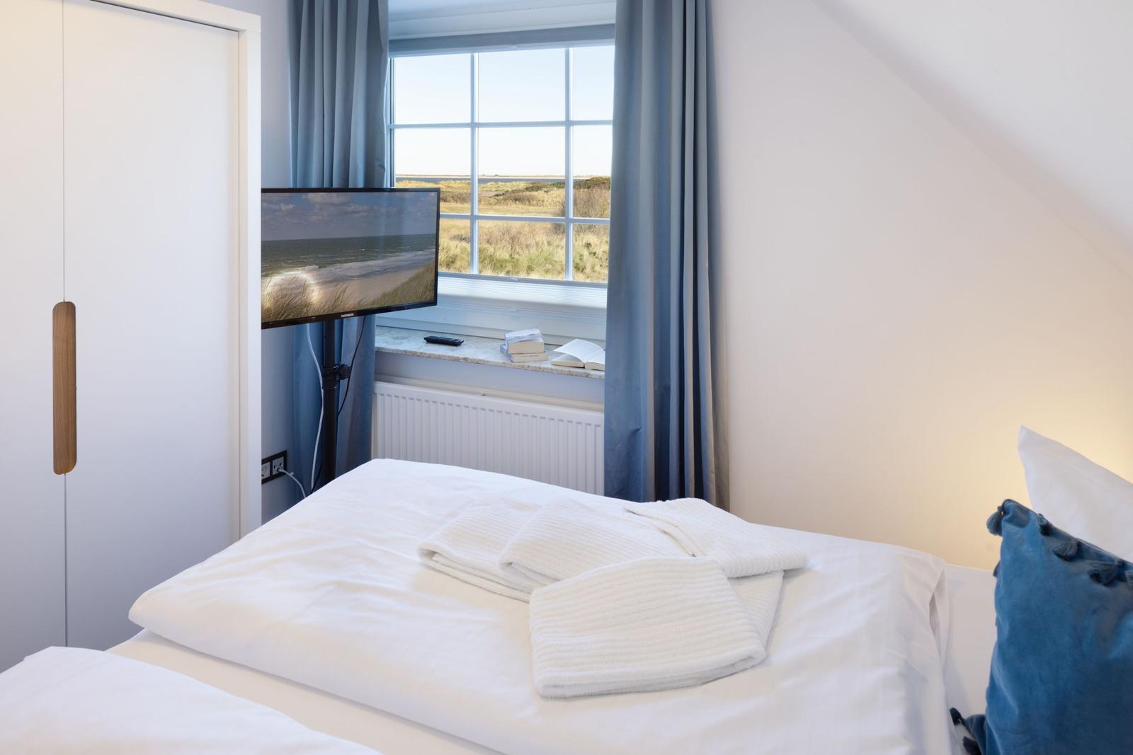 Schlafzimmer mit Bett, Fernseher und Fensterblick auf Dünen.
