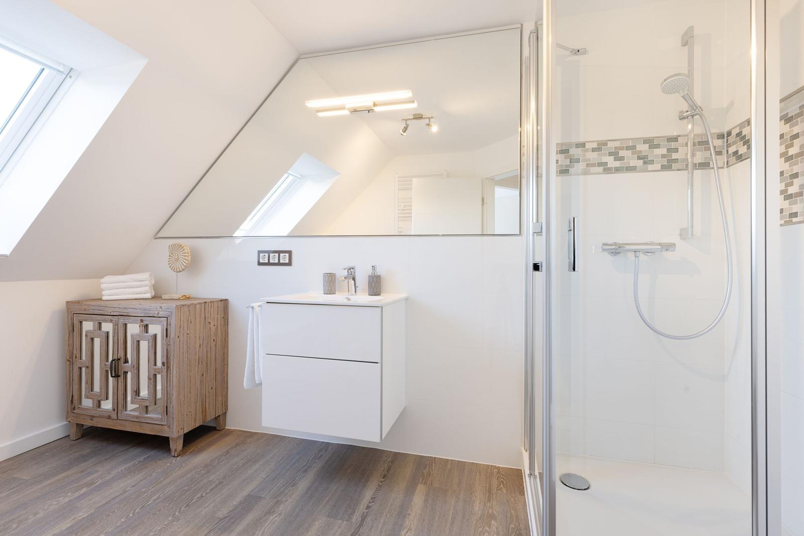 Badezimmer mit Dusche, Waschbecken und Holzregal unter Dachschrägen.