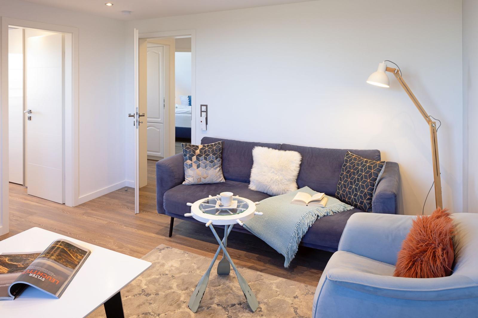 Wohnzimmer mit Sofa, Sessel und Tisch. Im Hintergrund sind Türen zu anderen Räumen.
