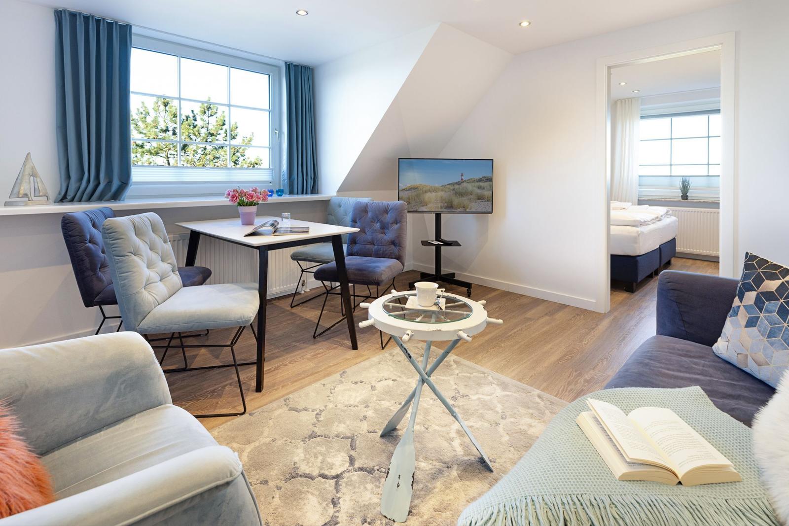 Wohnbereich mit Sofa, Tisch und Stühlen, Fernseher und Blick auf Schlafzimmer.