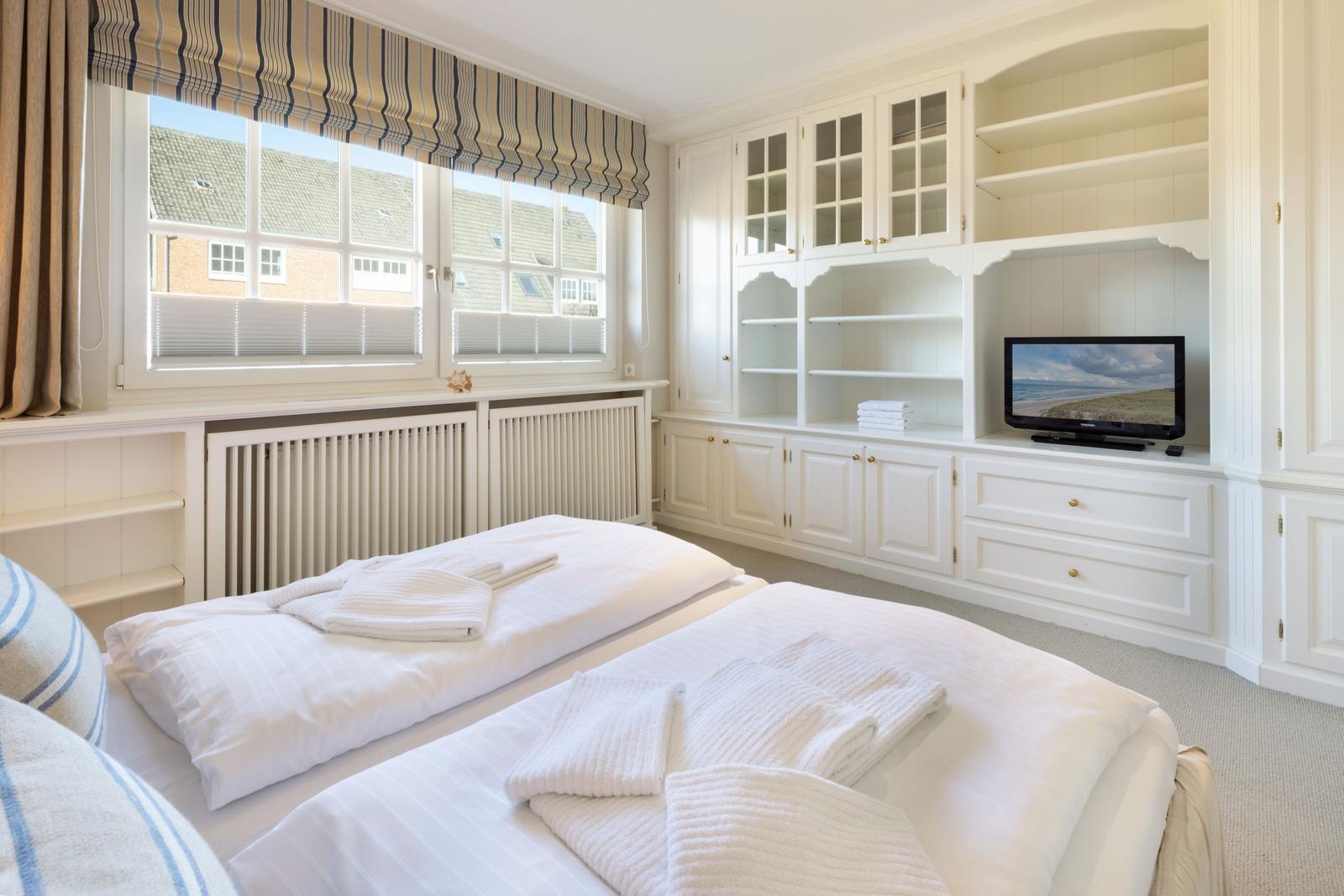 Schlafzimmer mit Doppelbett, weißen Schränken und TV. Fenster mit Rollladen und Blick auf Dächer.