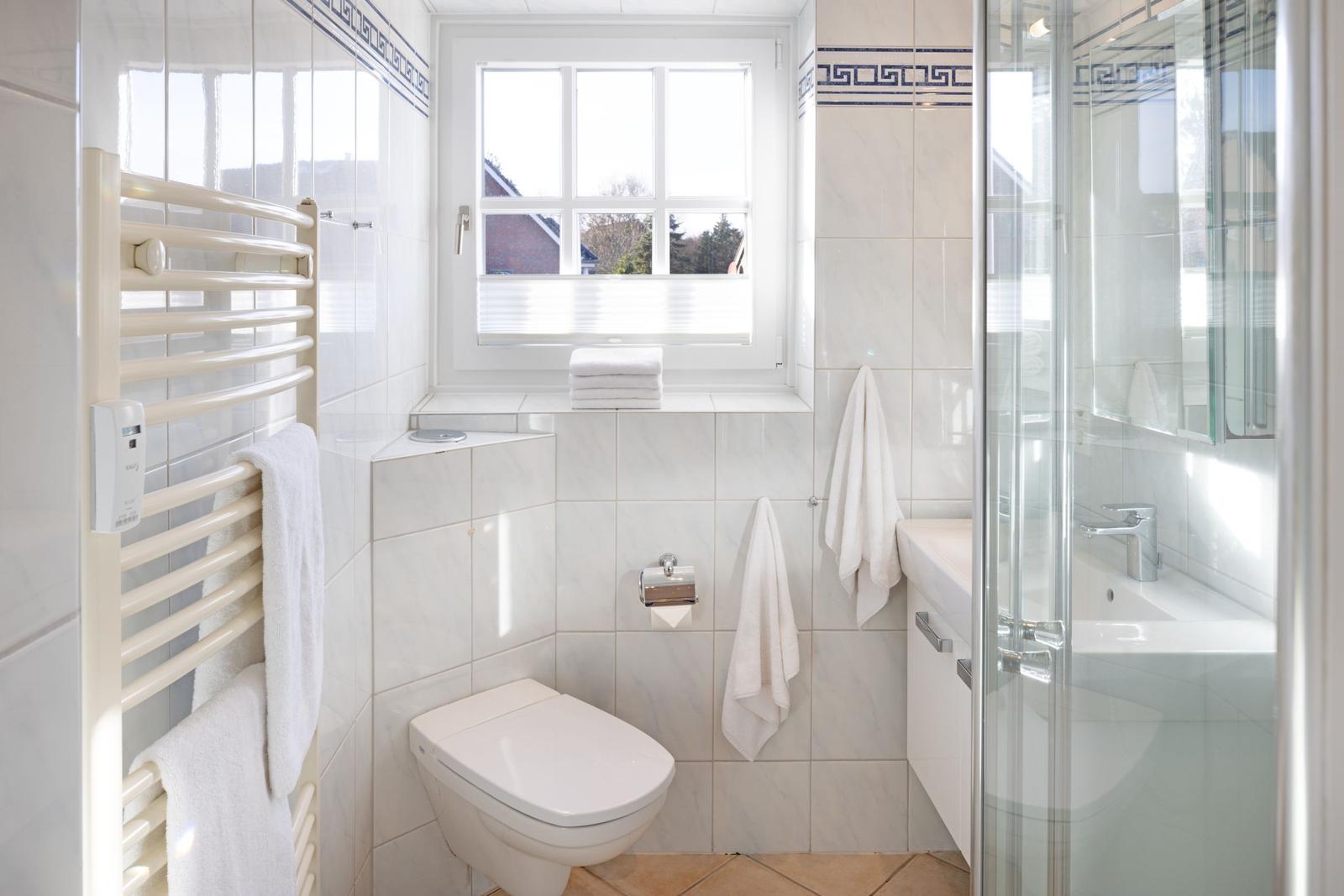 Badezimmer mit Dusche, Toilette und Fenster. Weiße Fliesen und Handtücher.