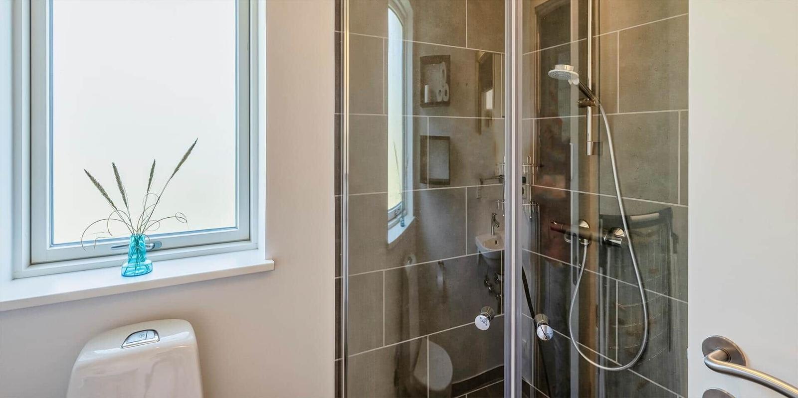 Bad mit Dusche, Fenster und Toilette. Auf dem Fensterbrett steht eine blaue Vase mit Pflanzen.
