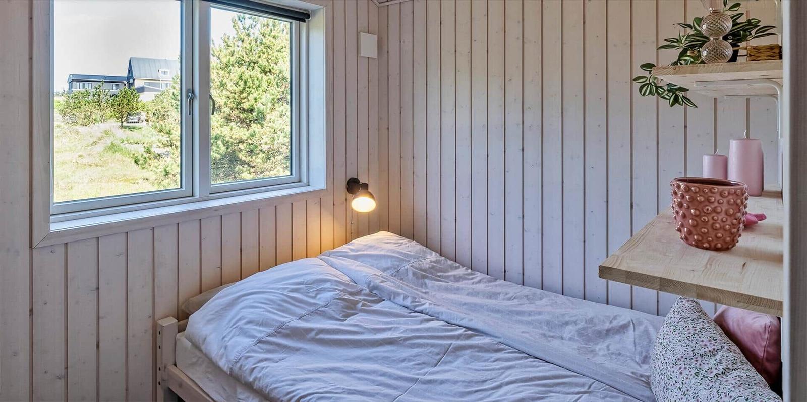 Ein Schlafzimmer mit Bett, Fenster und Holzwand. Auf Regal stehen Pflanzen und Kerzen.