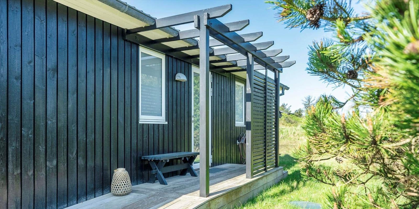 Schwarzes Holzhaus mit überdachter Terrasse und Bänken. Grüne Nadelbäume im Vordergrund.