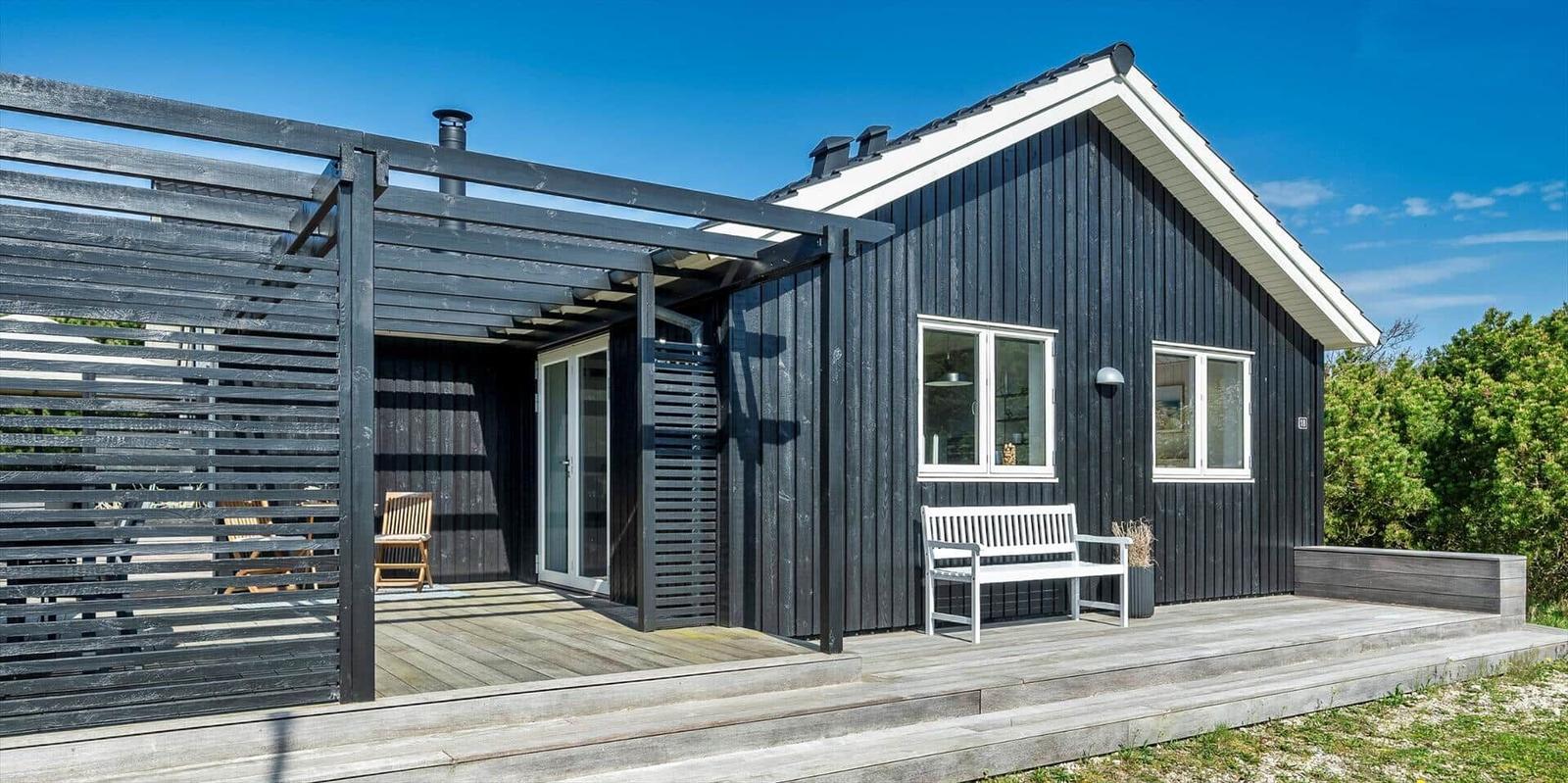Schwarzes Holzhaus mit Terrasse, weiße Bank und Sitzgelegenheit unter Pavillon.
