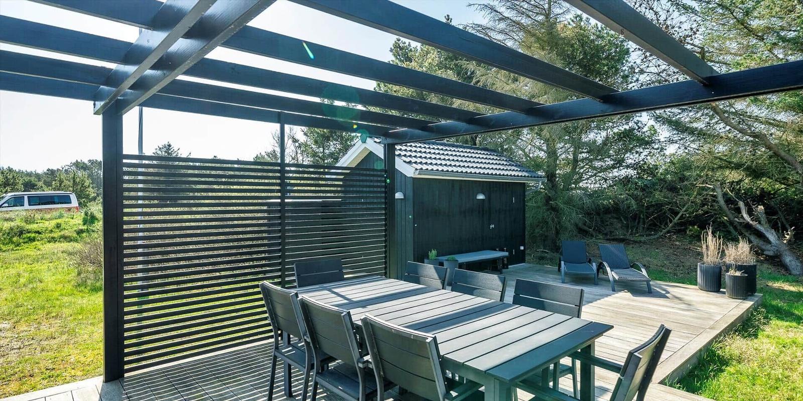 Terrasse mit Tisch, Stühlen und Sonnenschutz unter Pergola. Hintergrund: Wald und kleines Gebäude.