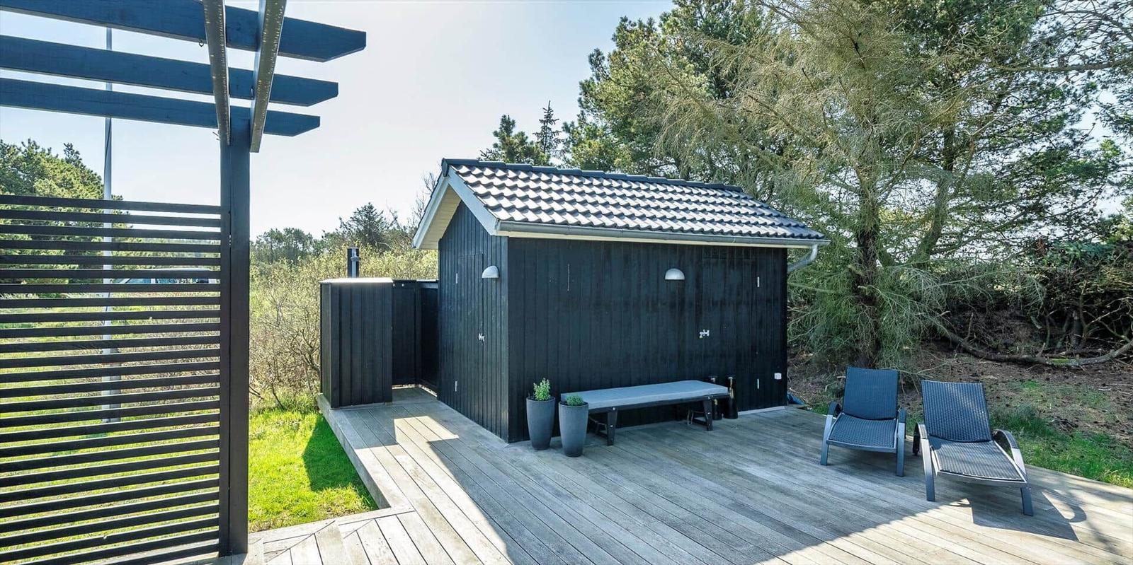 Schwarze Sauna mit Holzterrasse, Liegestühle und Pflanzen im Garten.