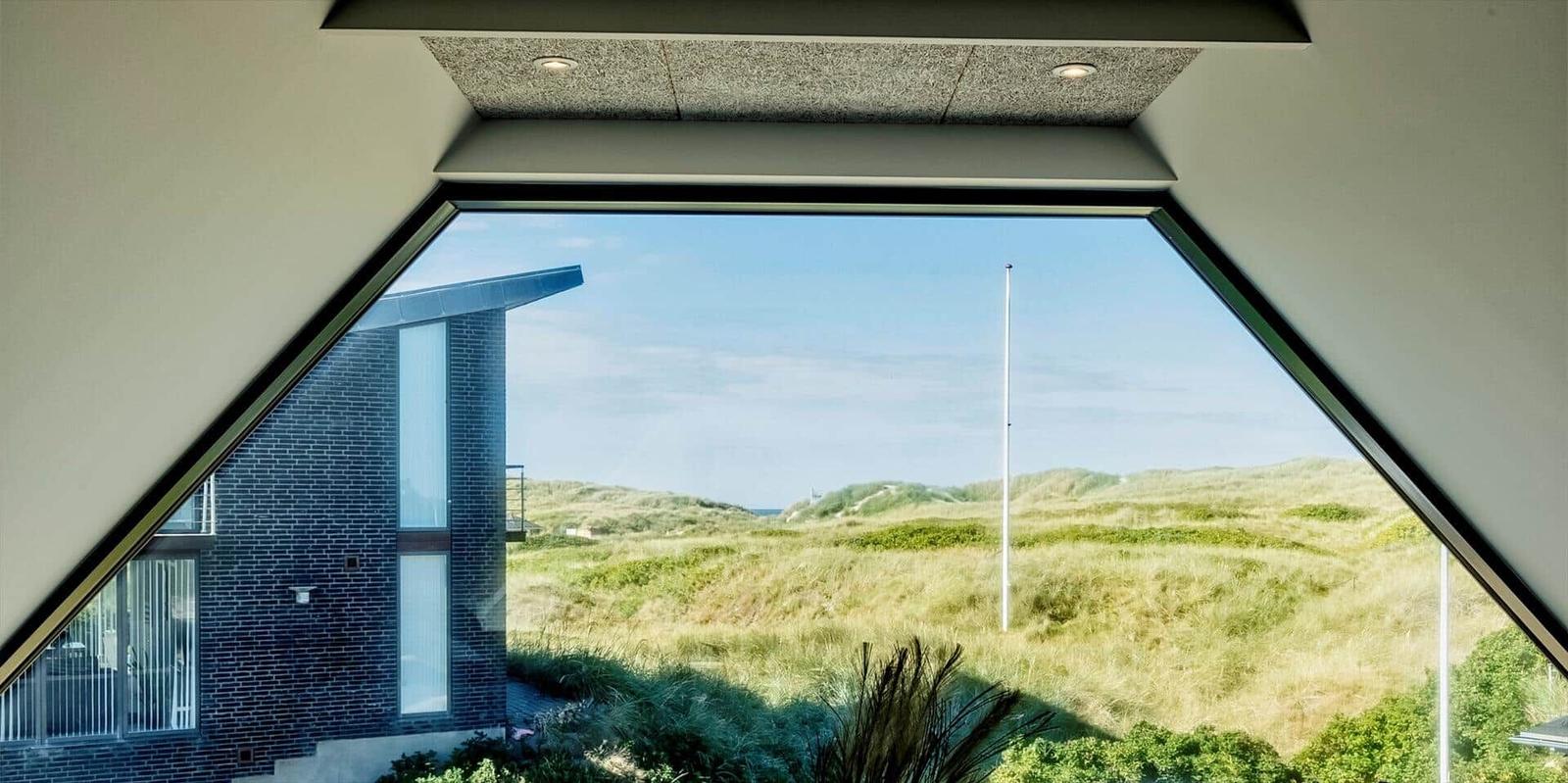 Durch ein großes Fenster blickt man auf Dünen und ein modernes Haus.