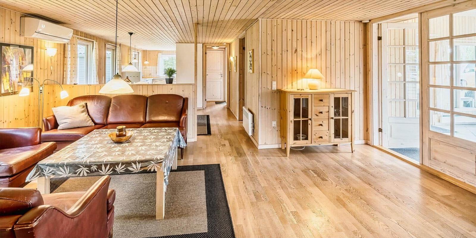 Wohnzimmer mit Holzwänden, Ledersofa und Holzboden. Durchgang zu Küche und Terrasse.