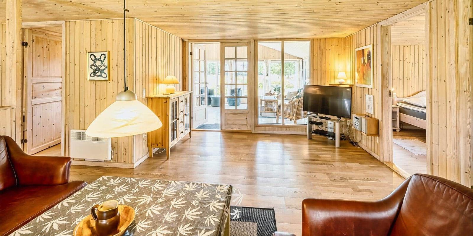 Wohnzimmer mit Holzwand, Fernseher und Sitzgelegenheiten. Durchgang zur Terrasse und Schlafzimmer.
