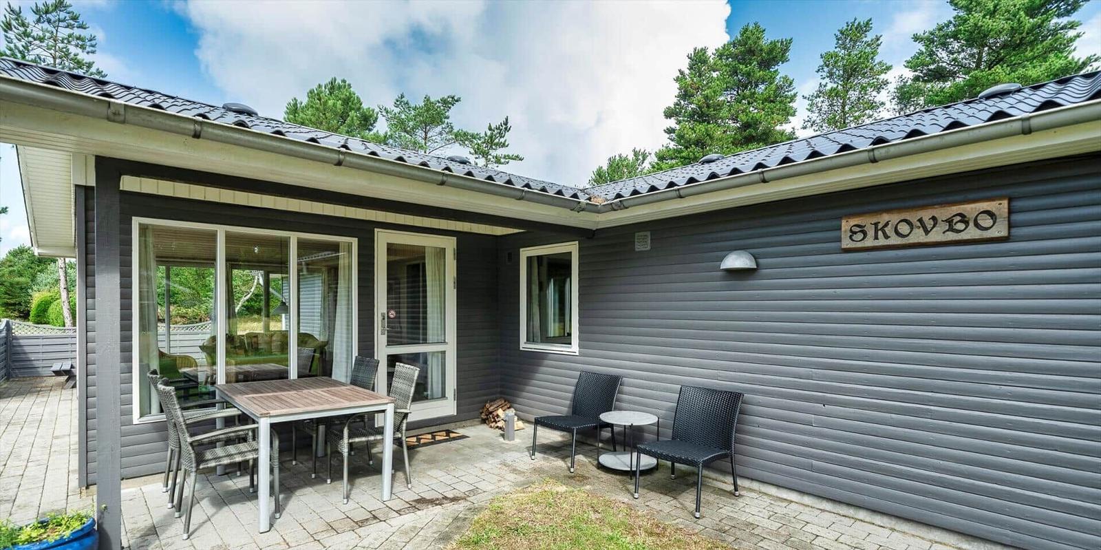 Haus mit Terrasse, Sitzgelegenheiten und Holzschild mit Namen. Umgeben von Bäumen und blauem Himmel.