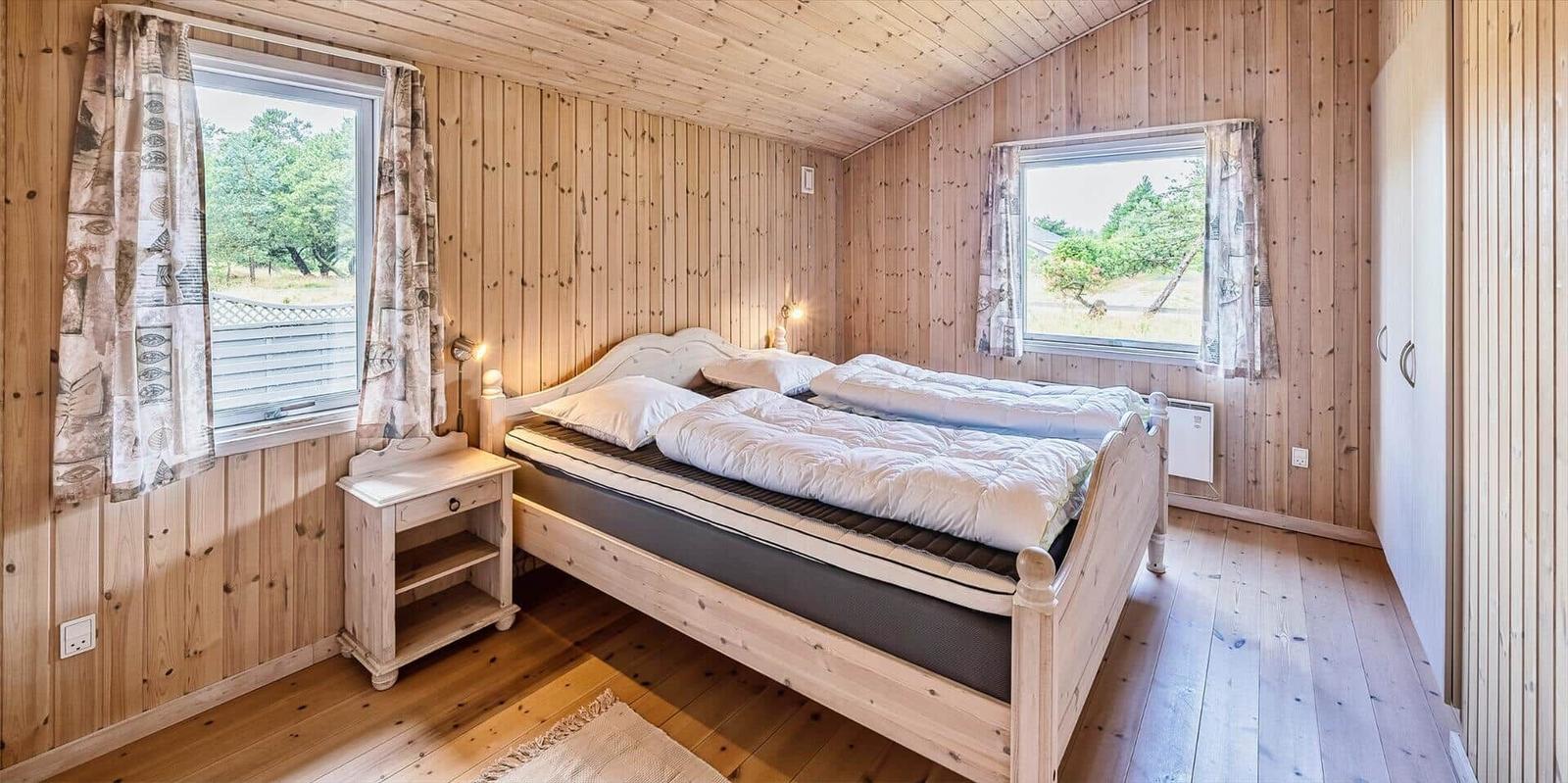 Schlafzimmer mit Holzwand, Bett, Nachttisch und zwei Fenstern.