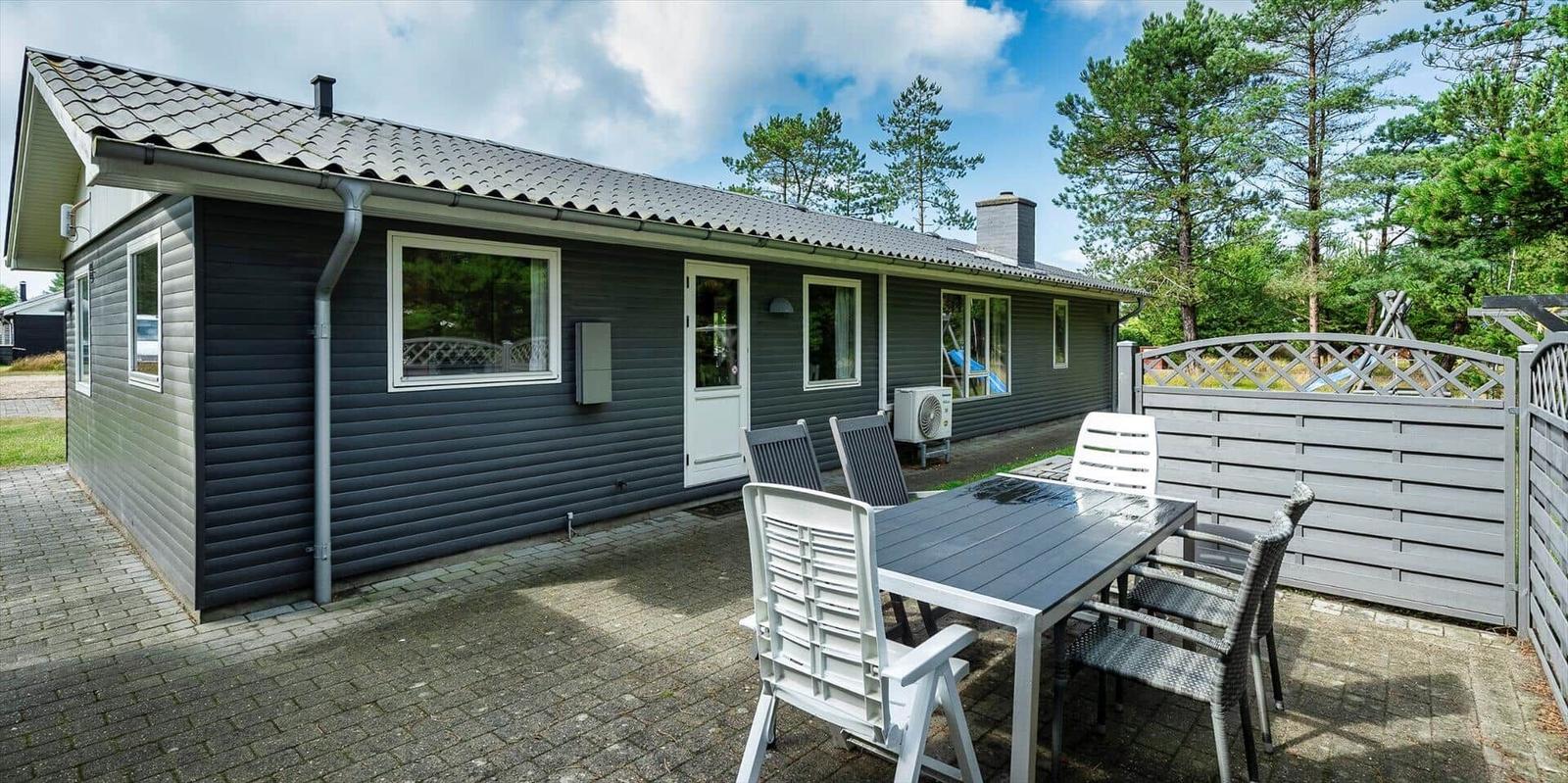 Ein graues Ferienhaus mit Terrasse und Gartenzaun. Gartenmöbel stehen auf dem gepflasterten Hof.