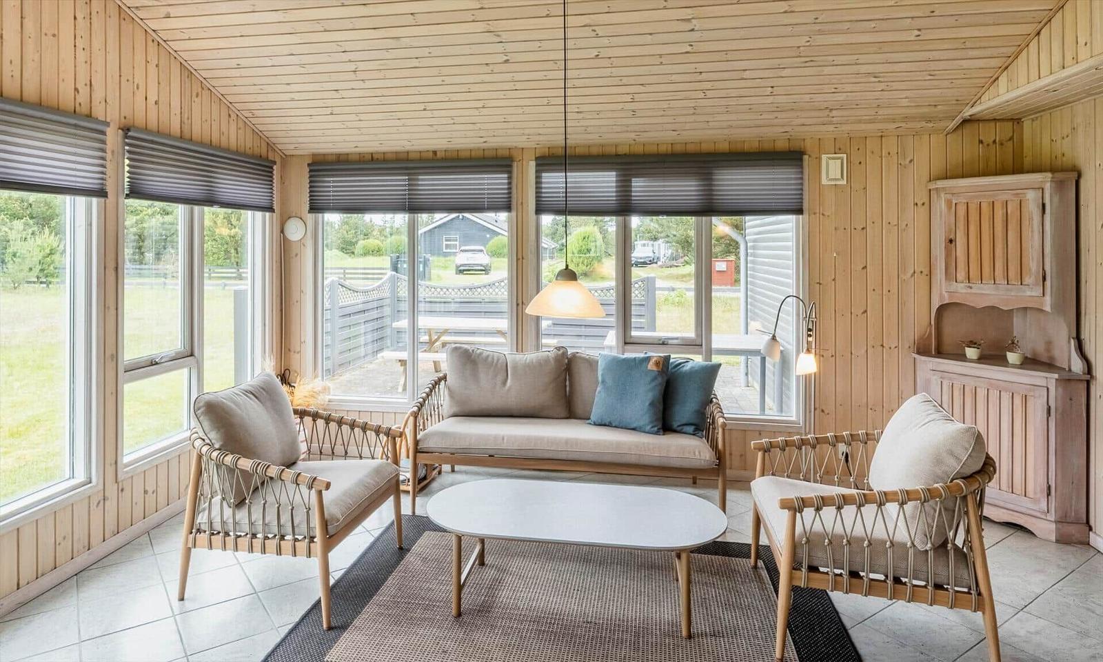 Wohnzimmer mit Holzwand, Sofa, zwei Sessel und Tisch. Große Fenster mit Blick nach draußen.