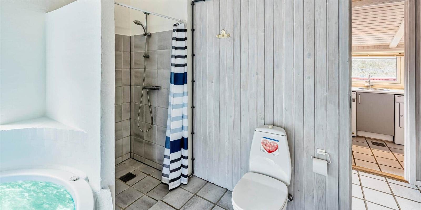 Badezimmer mit Dusche, Toilette und Whirlpool. Holzwand und Blick in die Küche.