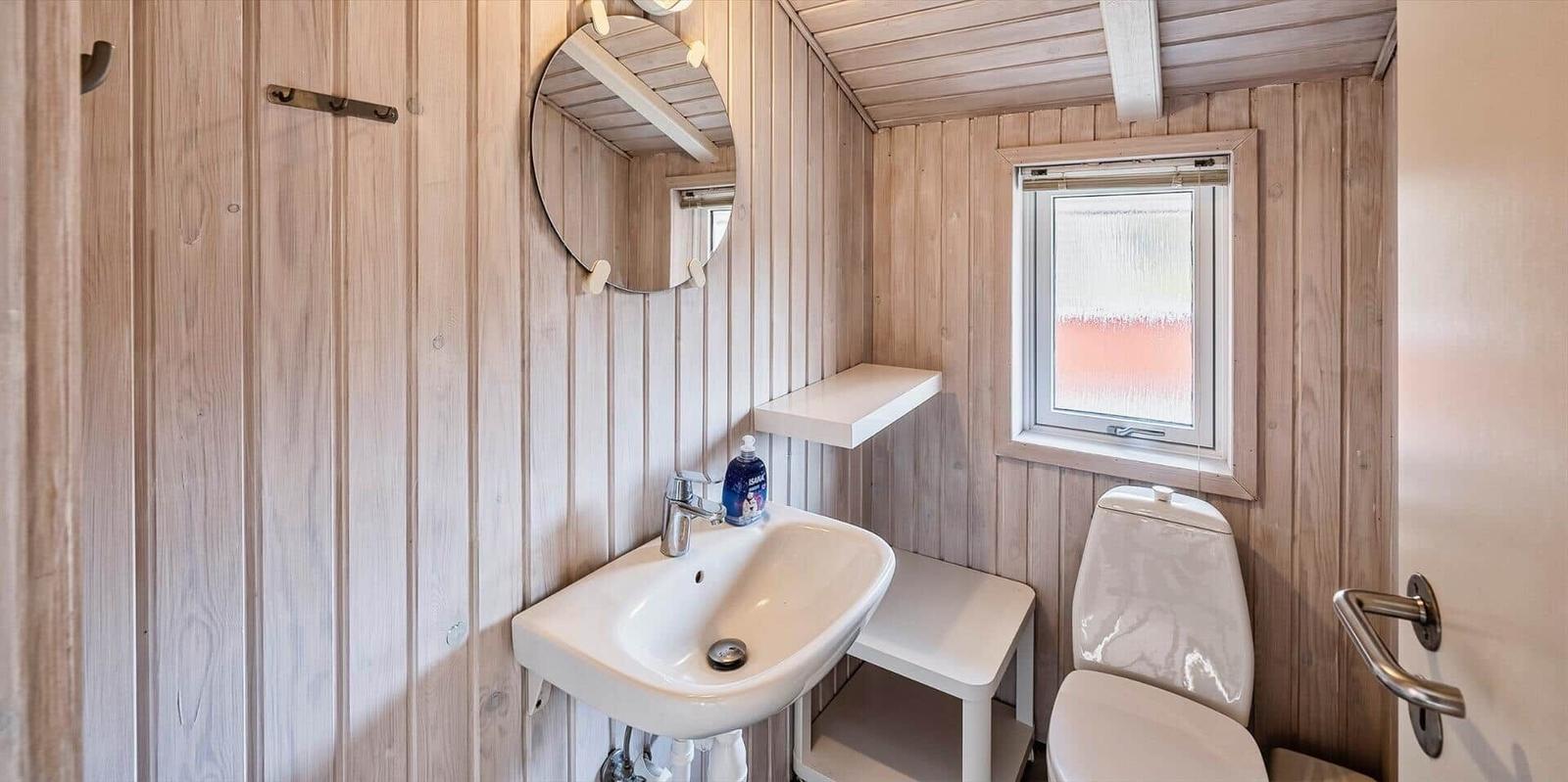 Badezimmer mit Waschbecken, Spiegel und Toilette. Holzwände und kleines Fenster.