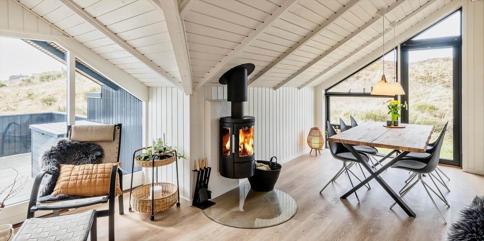 Ein Raum mit Kamin, Esstisch und großen Fenstern. Holzdielen und weiße Wände.
