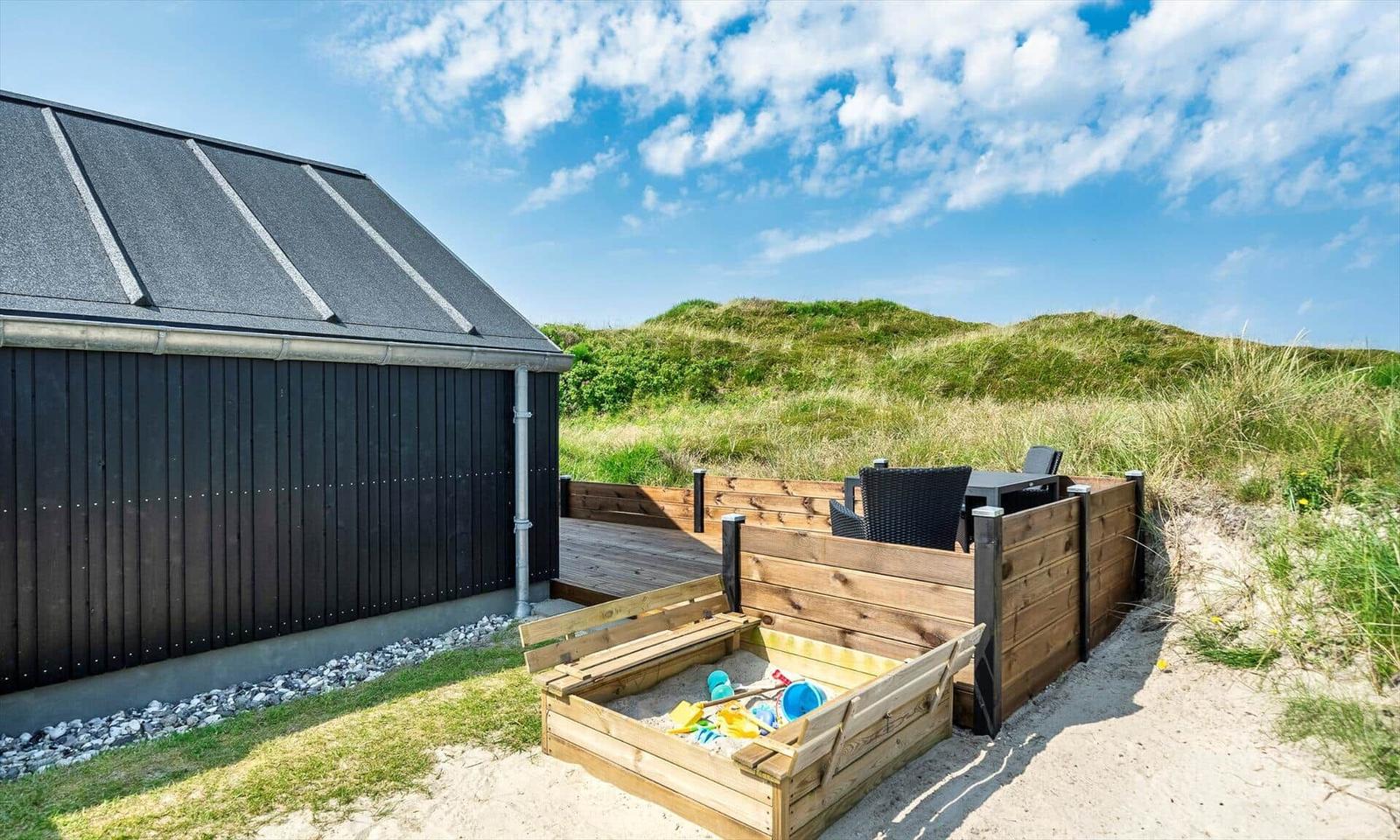 Holzterrasse mit Sitzgelegenheit und Sandkasten neben einem Haus mit dunkler Fassade.