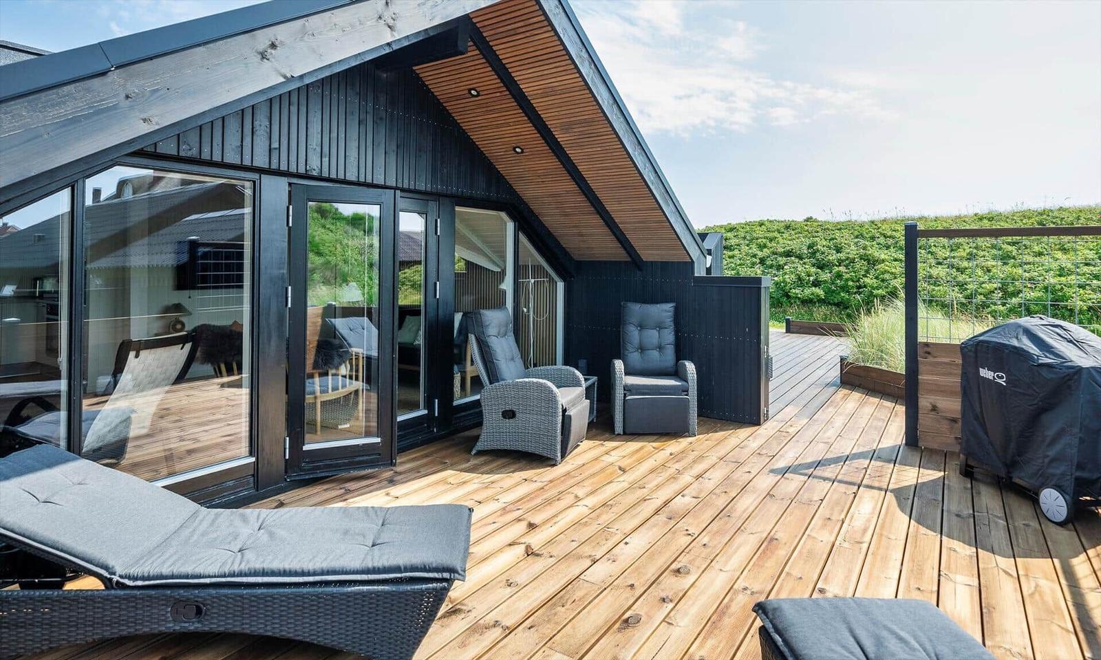 Terrasse mit Liegen und Sitzgelegenheiten. Holzdeck und Glaswände. Grill und Gartenzaun.
