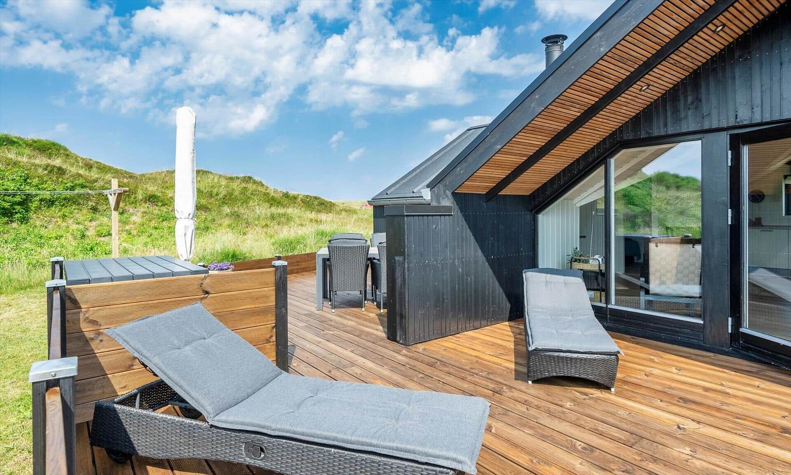 Terrasse mit Liegen, Tisch und Stühlen. Blick auf grüne Hügel. Modernes Haus mit Glasfront.
