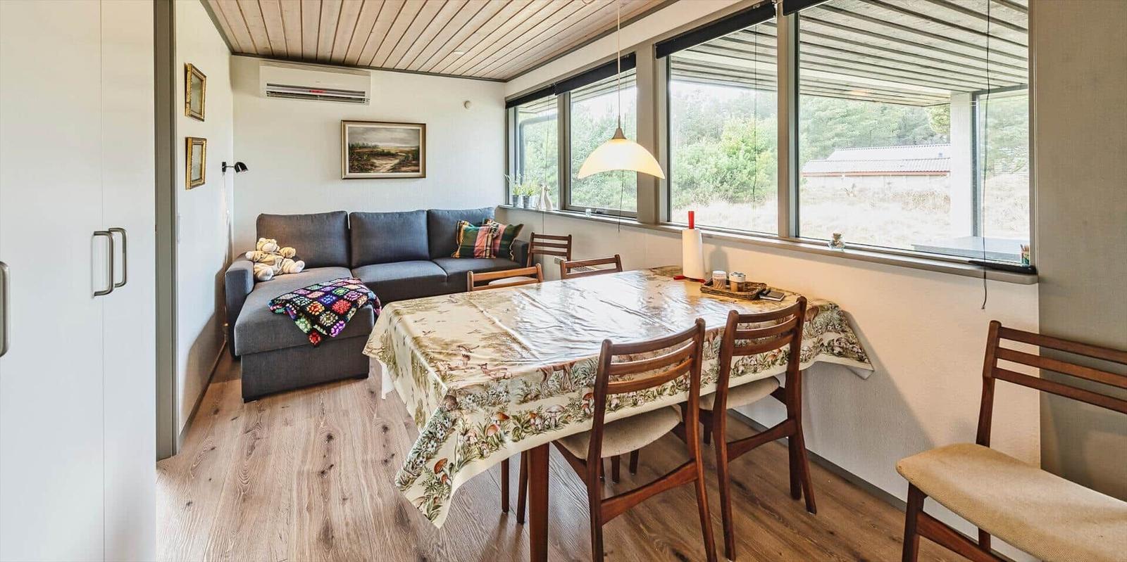 Wohn- und Essbereich mit Sofa, Tisch und Stühlen. Große Fenster mit Blick auf die Landschaft.