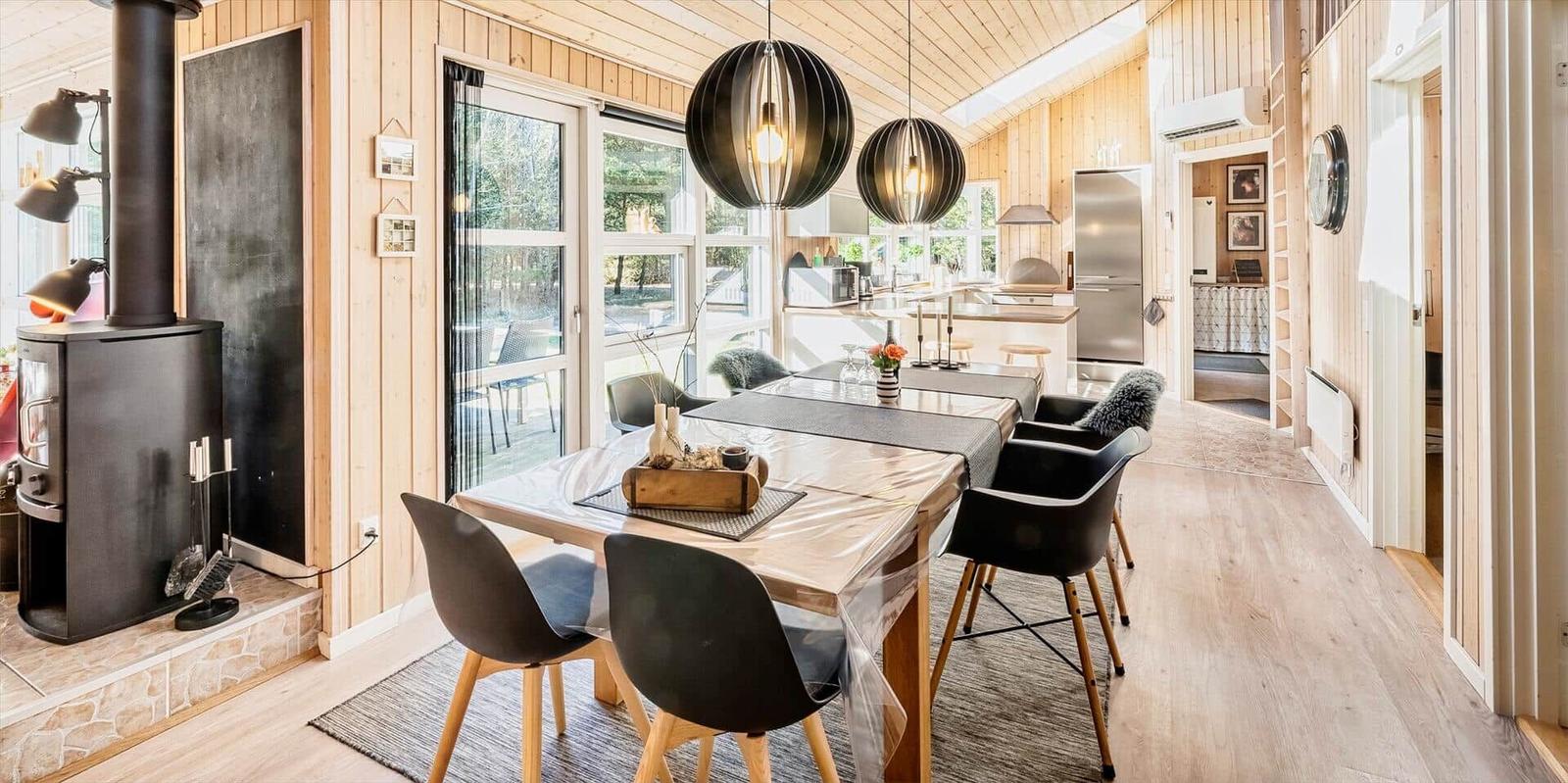 Essbereich mit Tisch, Stühlen und Kaminofen. Küche im Hintergrund. Holzwände und Boden.