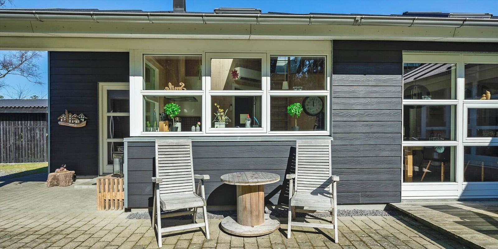 Haus mit Terrasse, zwei Stühlen und Tisch vor Fenstern mit Blick ins Innere.