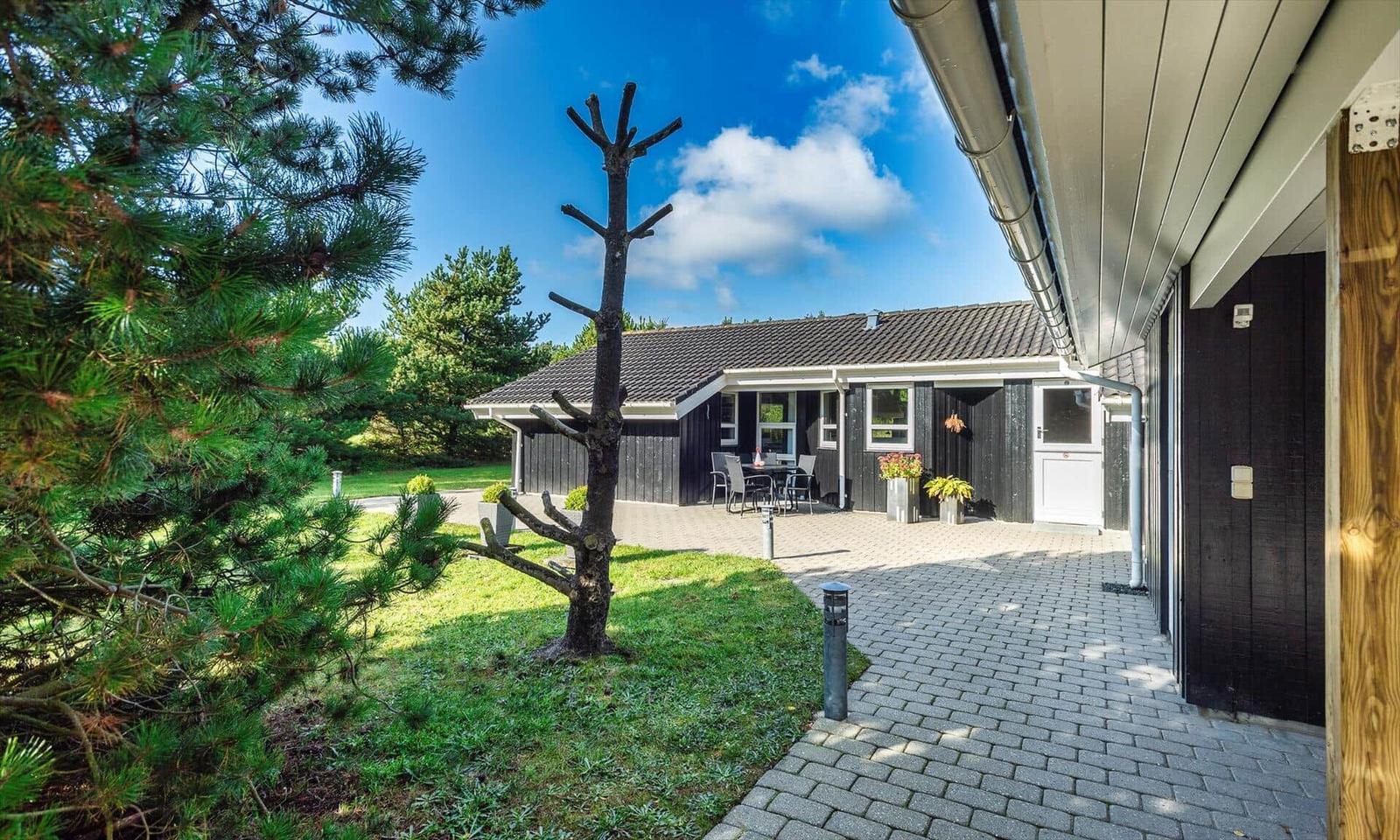 Schwarzes Holzhaus mit Terrasse, Garten und Bäumen unter blauem Himmel.
