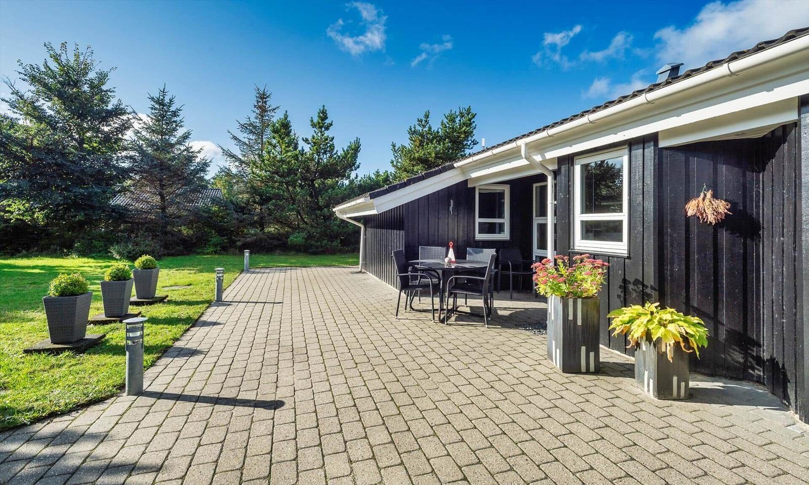 Schwarze Holzhütte mit Terrasse, Garten und Sitzgruppe unter blauem Himmel.