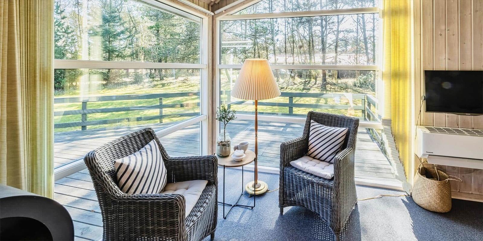 Zwei Rattan-Stühle mit Kissen stehen vor großen Fenstern mit Blick auf eine Terrasse und Wald.