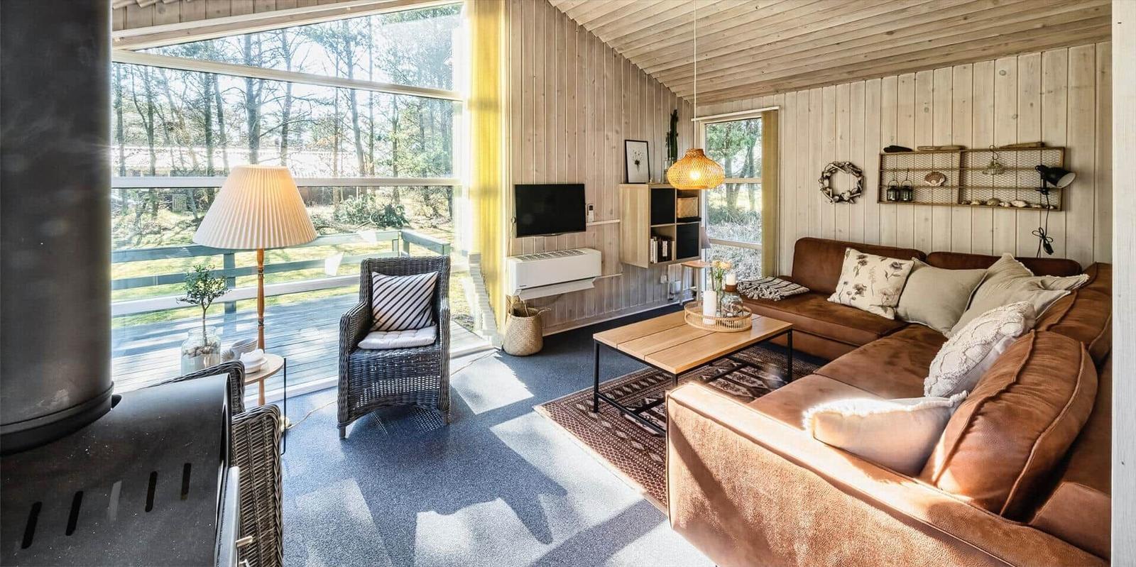 Wohnzimmer mit Holzwänden, Sofa, Couchtisch und Fernseher. Große Fenster mit Blick auf den Garten.