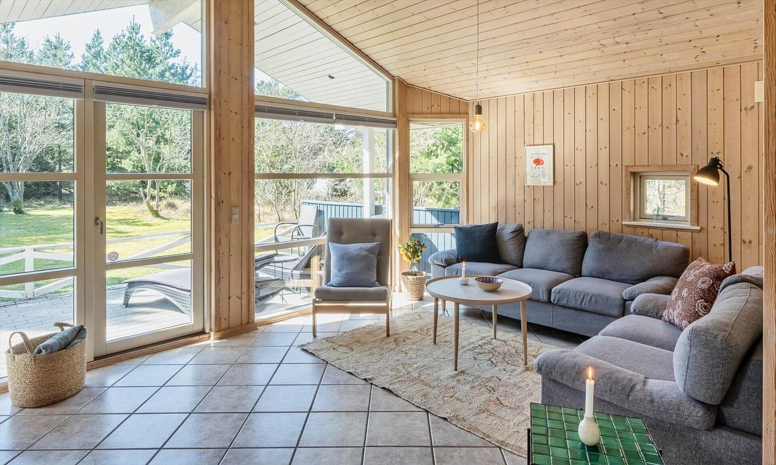 Wohnraum mit Holzwänden, Sofa, Sessel und großem Fenster zum Garten.