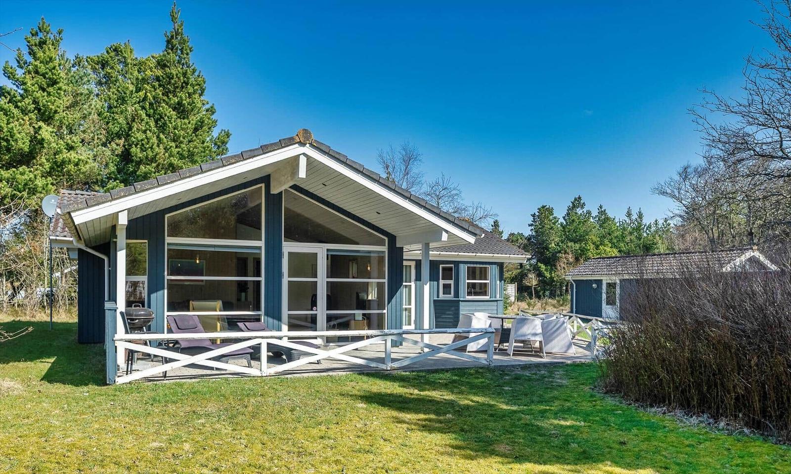 Ein Haus mit Terrasse, Grill und Garten. Im Hintergrund sind Bäume und ein kleines Nebengebäude zu sehen.
