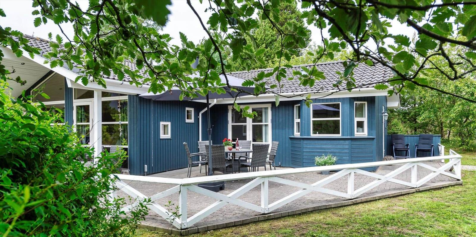 Blauer Holzhaus mit Terrasse und Garten. Tisch, Stühle und Sonnenschirm sind draußen.
