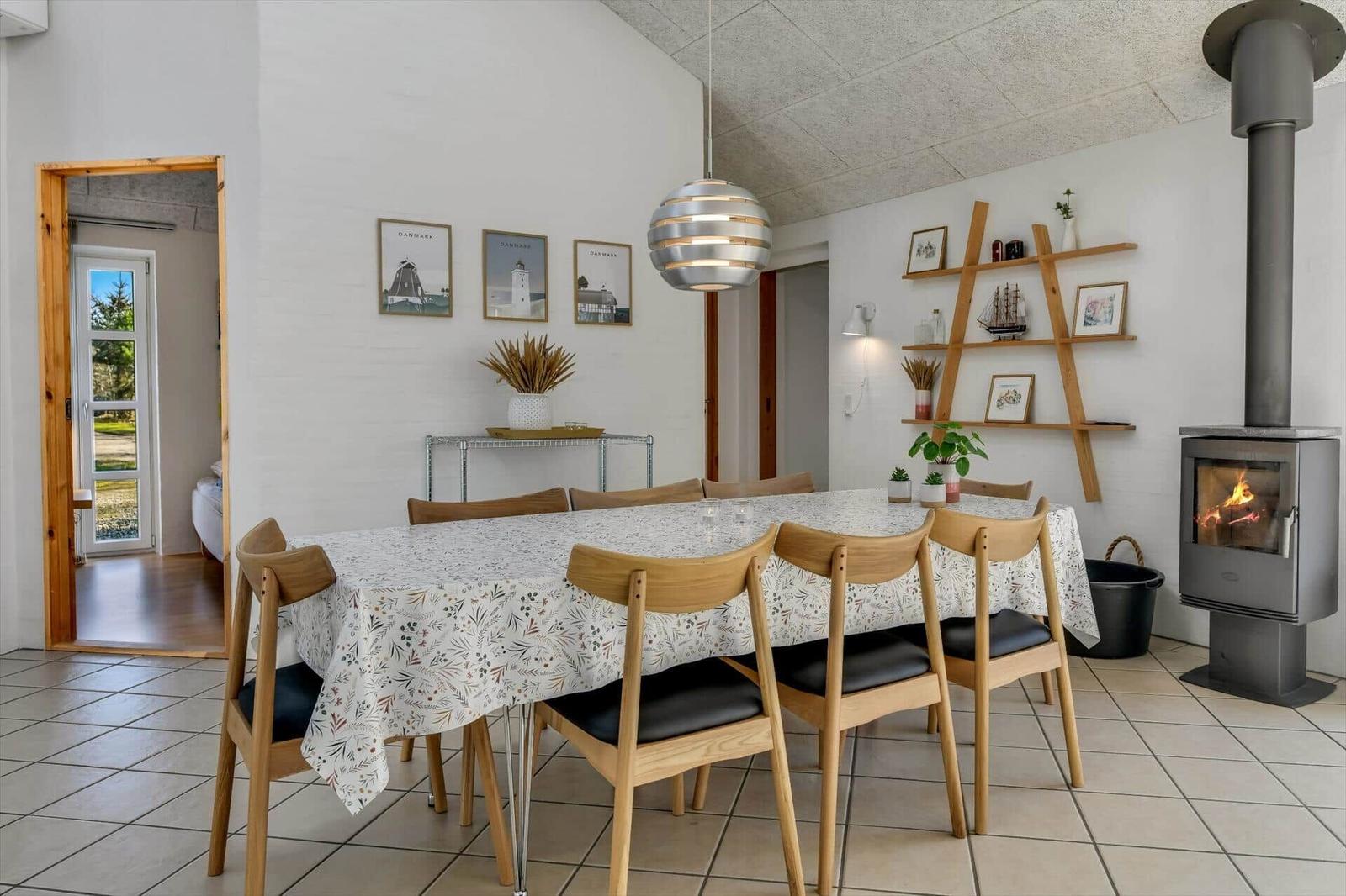 Esszimmer mit Holzstühlen, Tischdecke und Kaminofen. Wandregal und Bilder an der Wand.