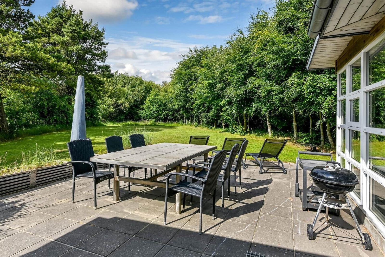 Terrasse mit Tisch, Stühlen und Grill neben Haus. Grünflächen und Bäume im Hintergrund.