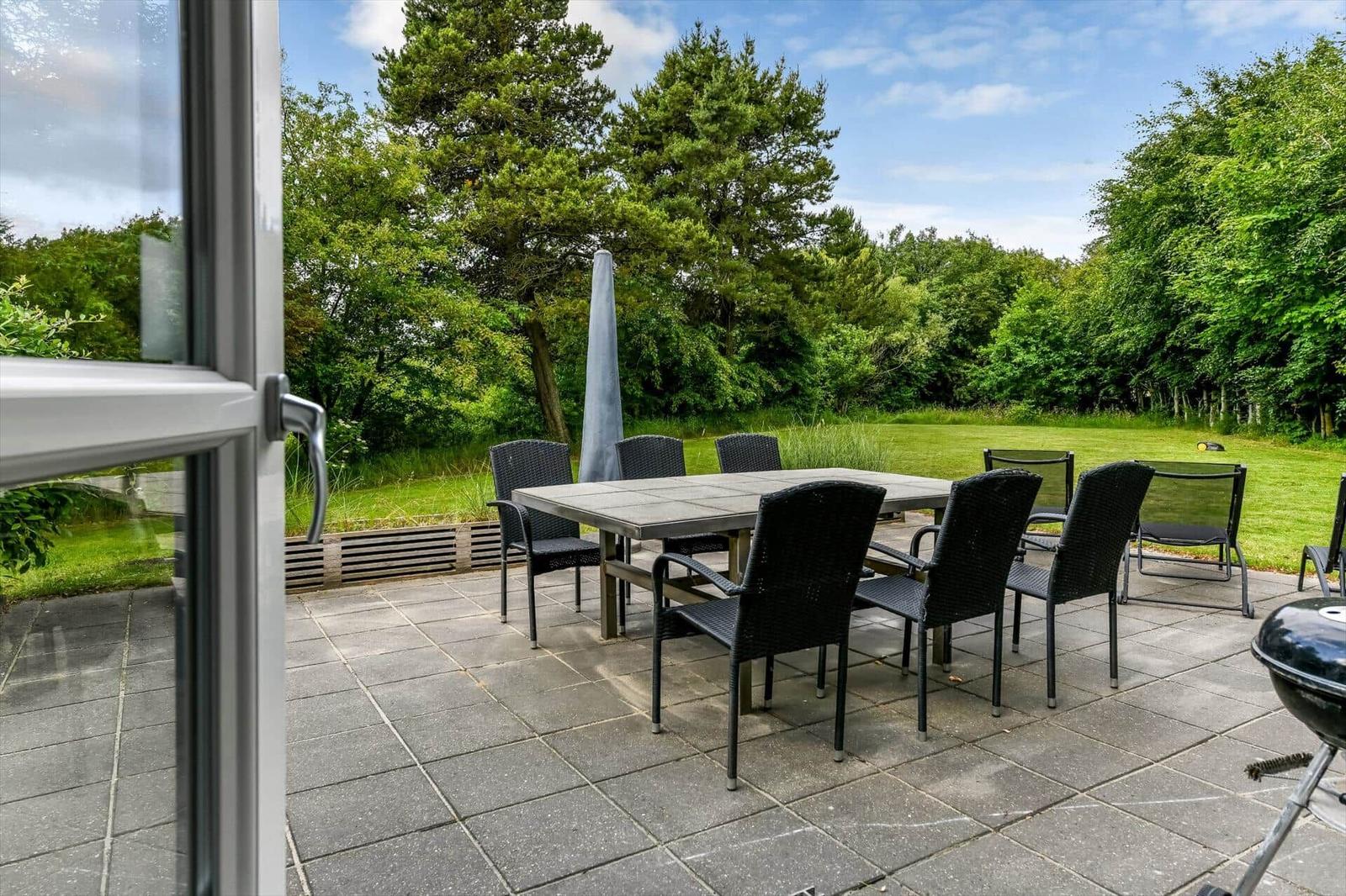 Terrasse mit Tisch, Stühlen und Grill. Grünfläche und Bäume im Hintergrund.