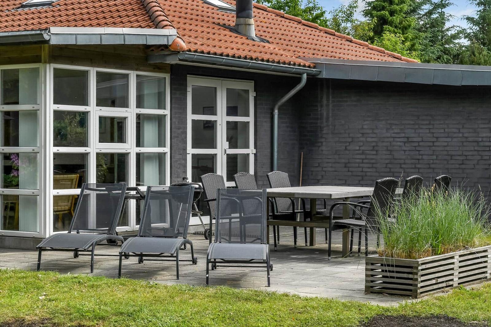 Terrasse mit Tisch, Stühlen und Liegestühlen vor einem Haus mit grauer Wand und rotem Dach.