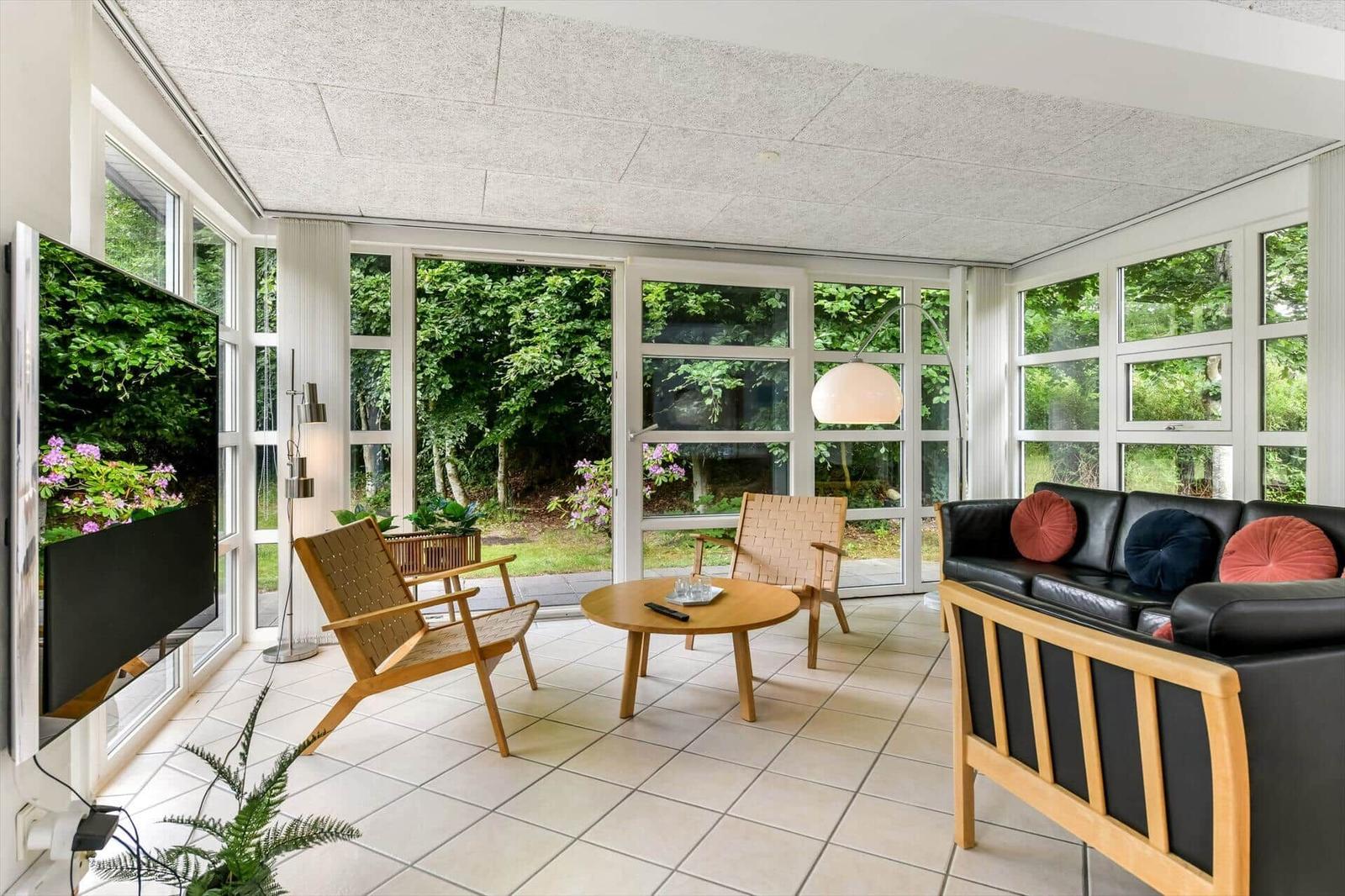 Ein Wohnzimmer mit Sofa, Stühlen und Fernseher. Große Fenster zeigen einen Garten.