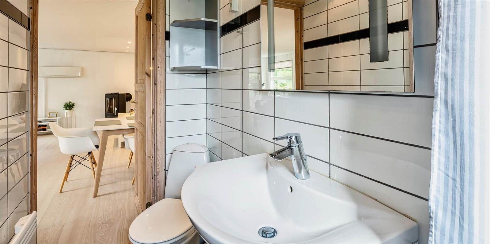 Badezimmer mit Waschbecken, Toilette und Spiegel. Durchgang ins Wohnzimmer mit Tisch und Stühlen.