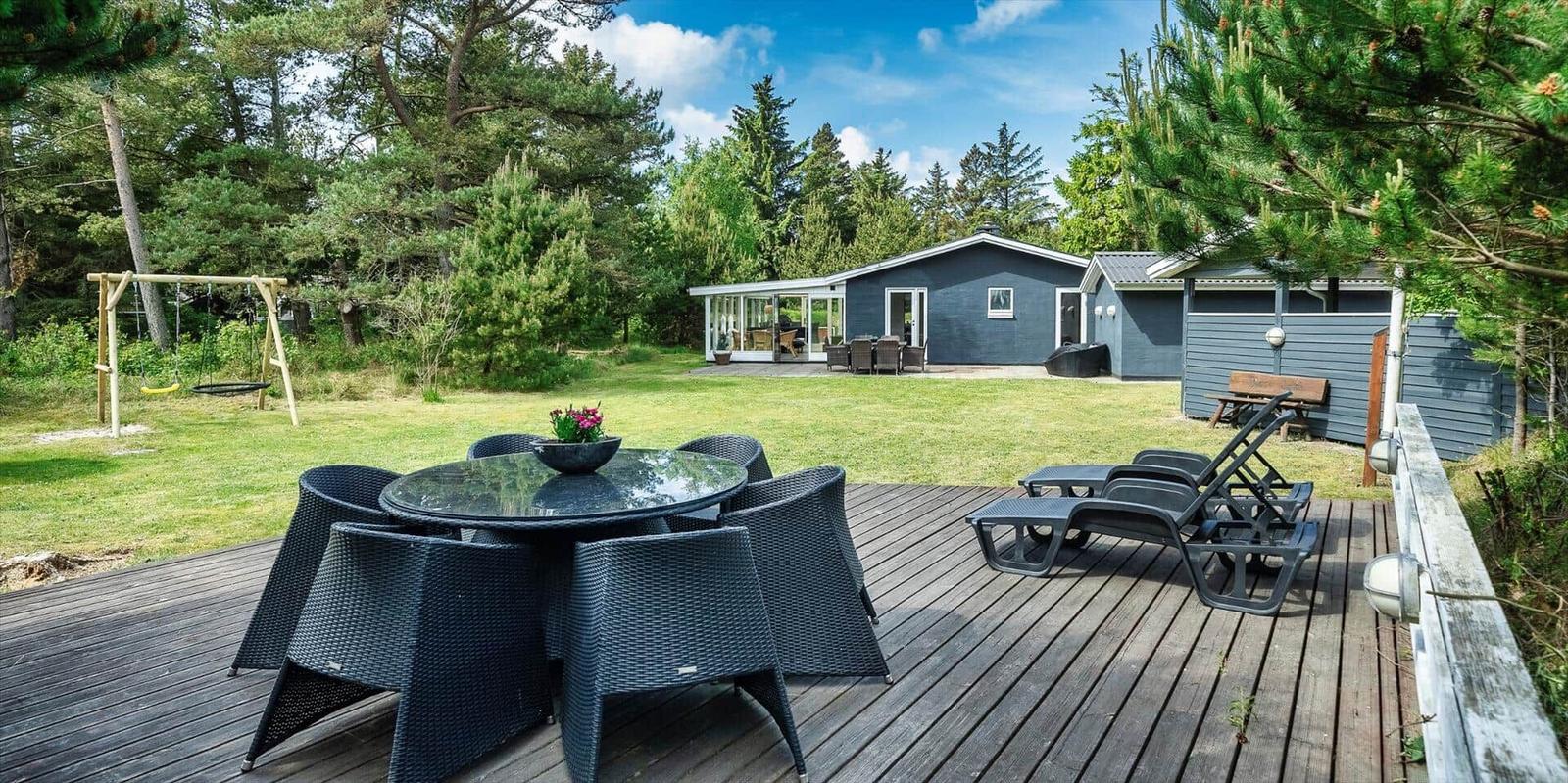 Terrasse mit Tisch, Stühlen und Liegen. Im Hintergrund Haus mit Garten und Spielplatz.