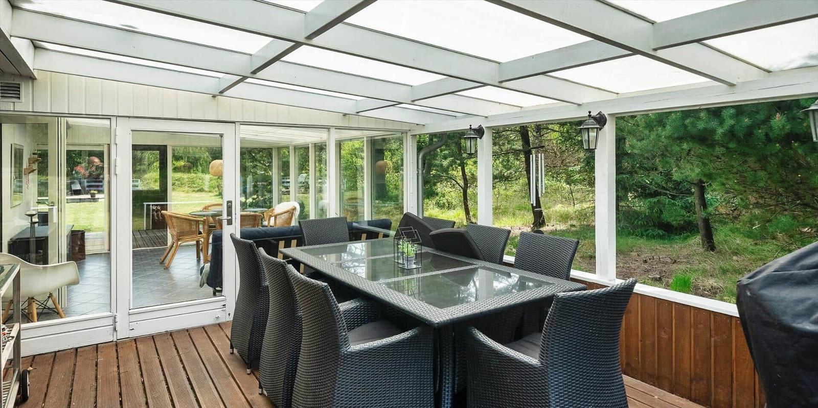Terrasse mit Glasdach, Esstisch und Stühlen. Hintergrund: grüne Bäume und Schiebetüren.
