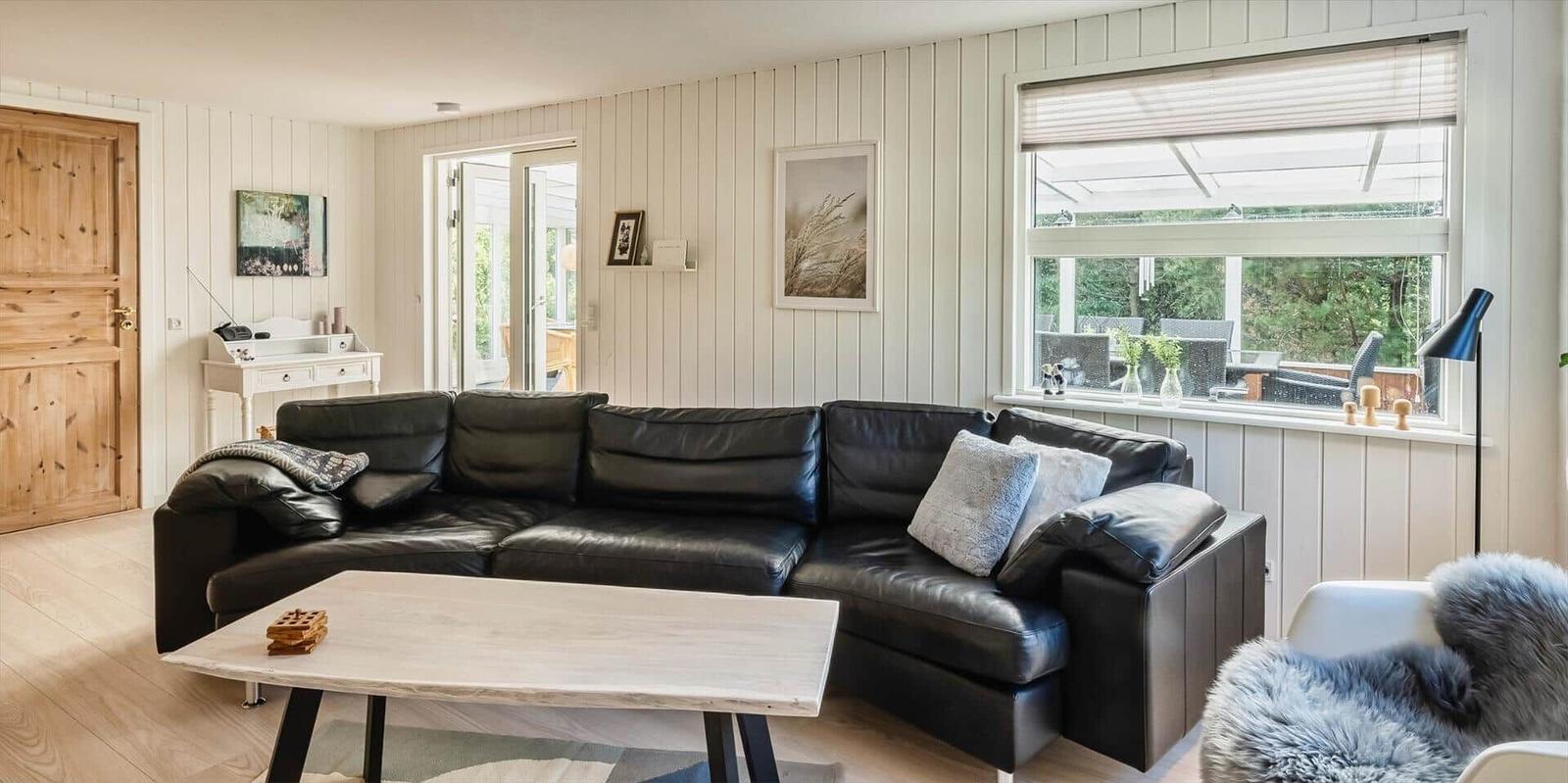 Wohnzimmer mit schwarzer Ledercouch, Holzboden und großem Fenster zur Terrasse.