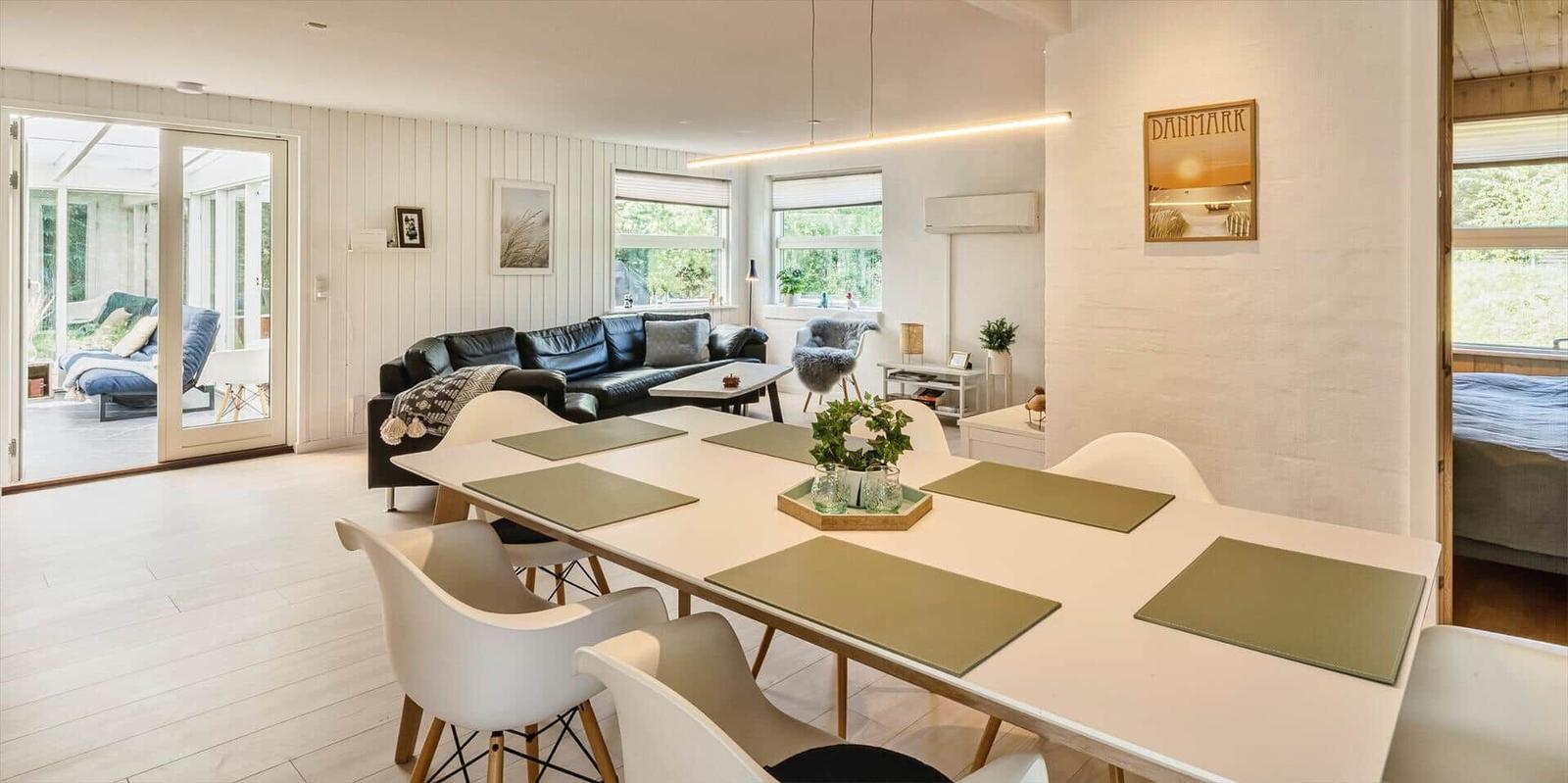 Essbereich mit Tisch und Stühlen. Im Hintergrund Wohnbereich mit Sofa und Fenstern.