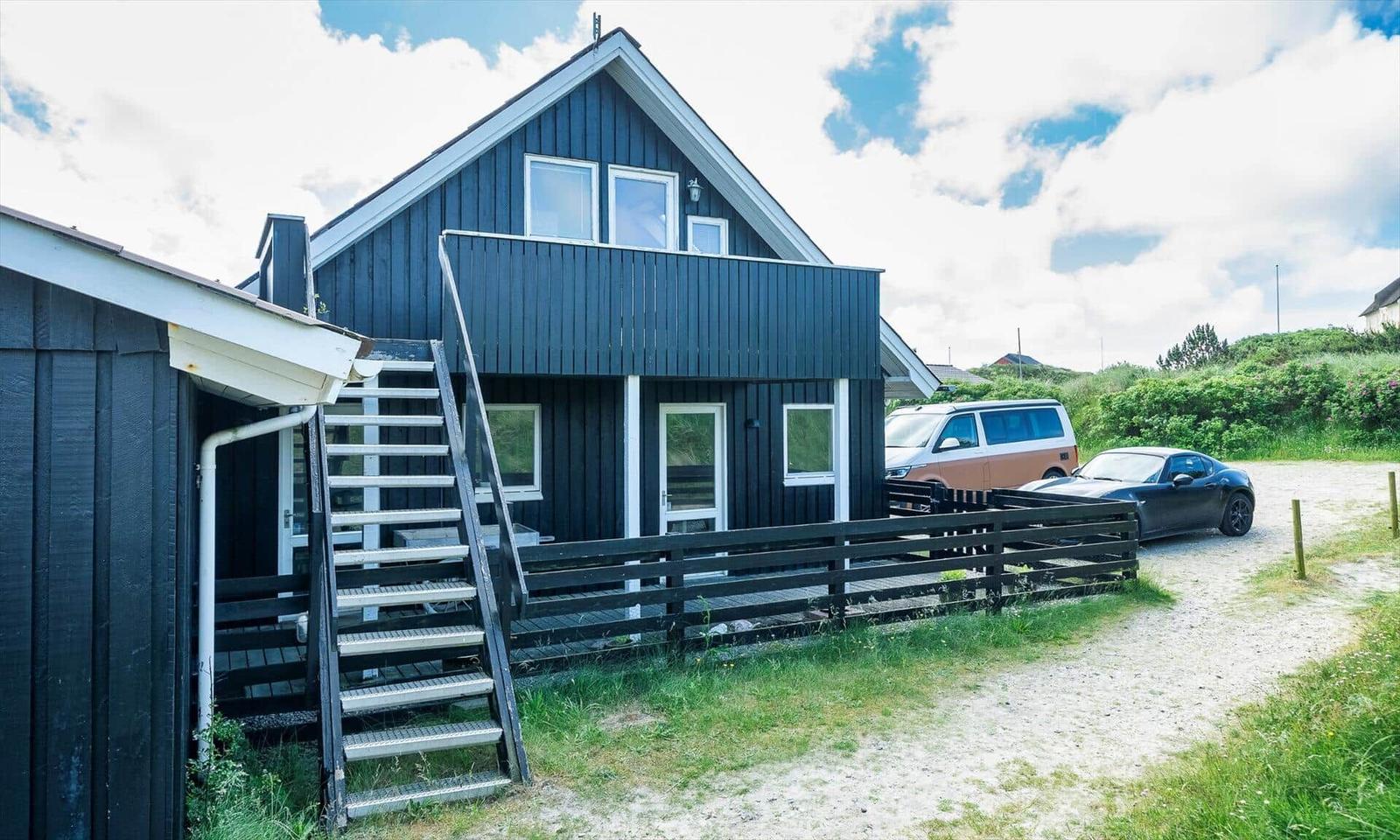 Schwarzes Holzhaus mit Balkon, Treppe und Parkplatz mit zwei Fahrzeugen.