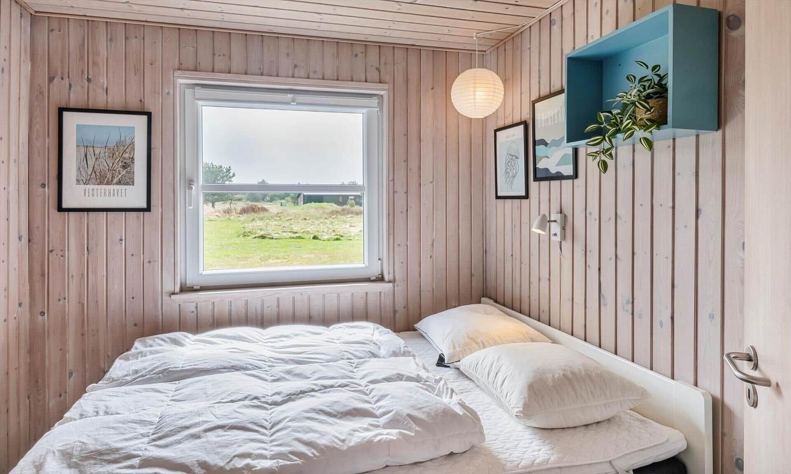 Schlafzimmer mit Holzwand, Fenster und Bett. An der Wand hängen Bilder und eine Pflanze.