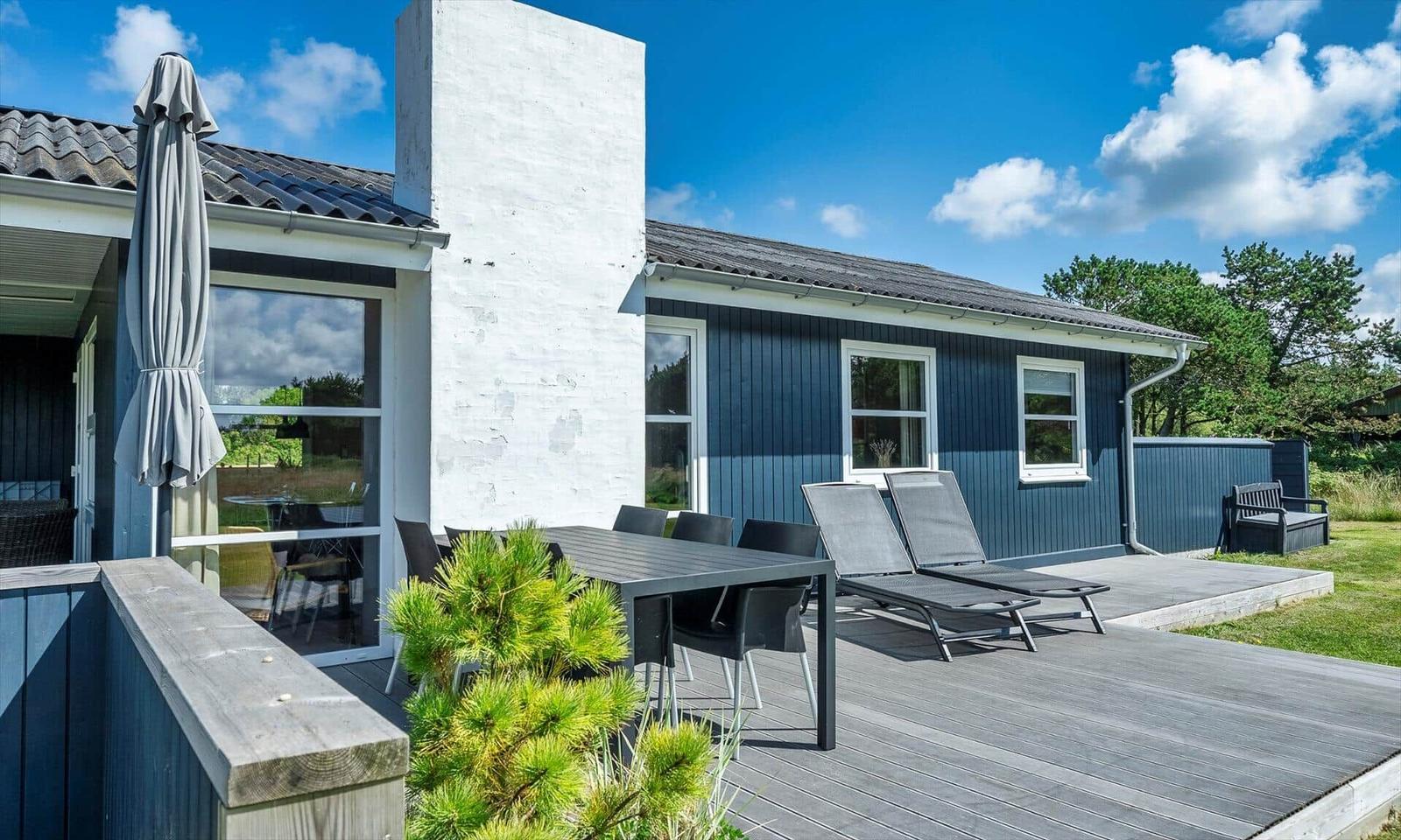 Terrasse mit Tisch, Stühlen und Liegen vor einem blauen Haus mit weißem Schornstein.