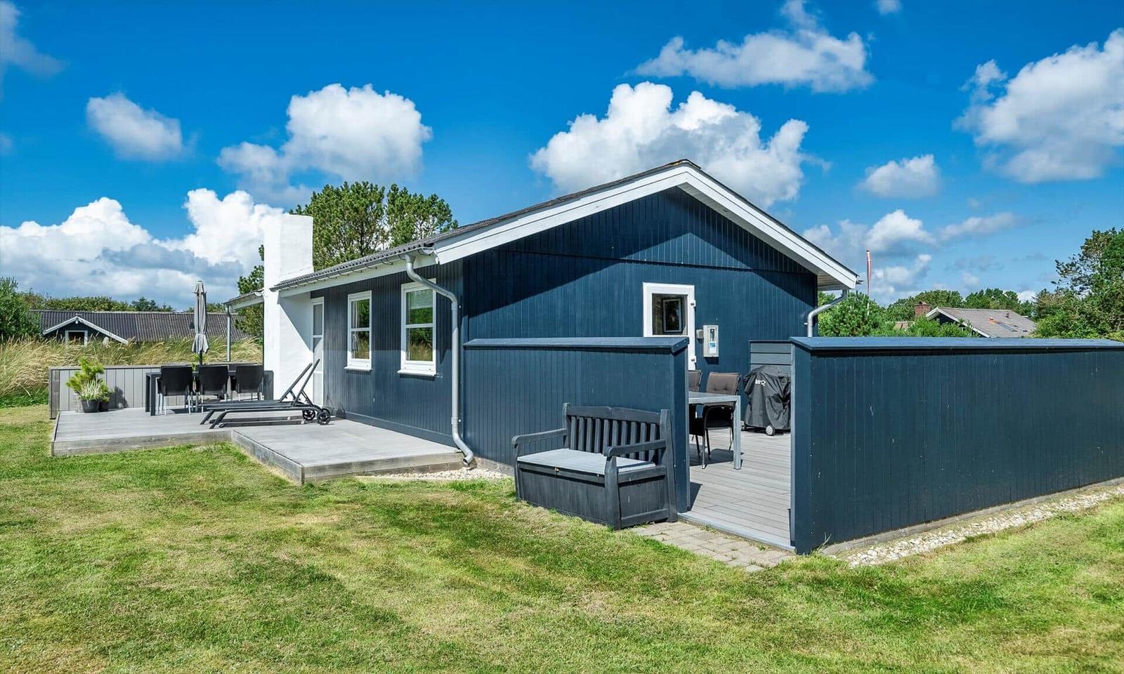 Ein dunkelblauer Ferienhaus mit Terrasse und Garten. Ein Gartenmöbel und Grill sind vorhanden.