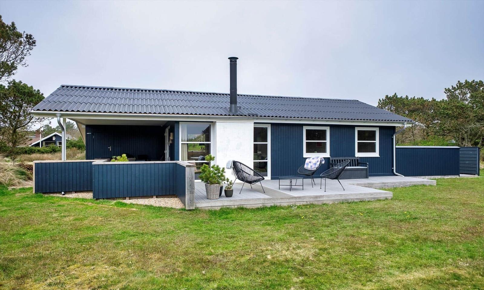 Ein blauer Holzhaus mit Terrasse und Garten. Drei Sitzgelegenheiten und ein Schornstein sind sichtbar.
